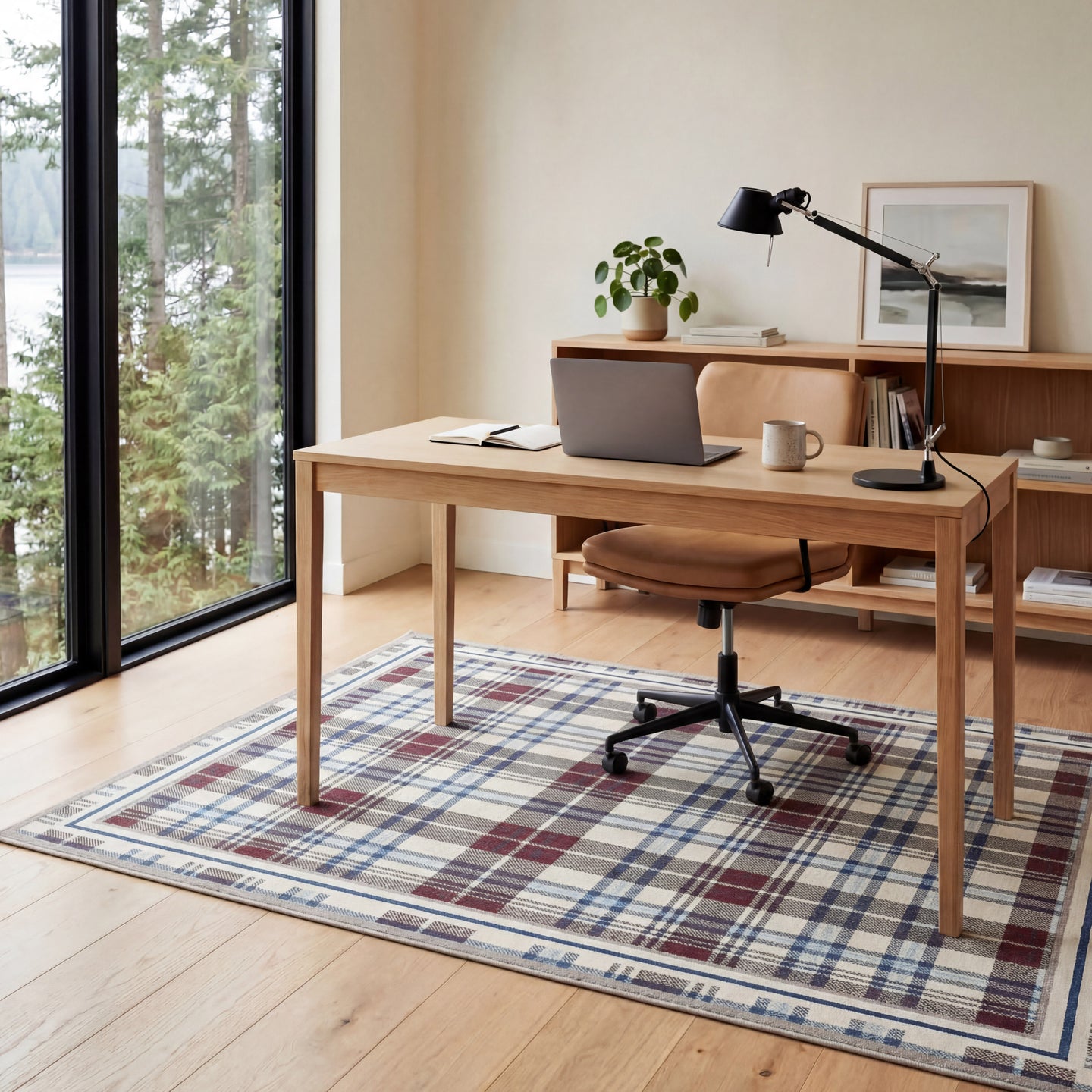 Home office with wooden desk, laptop, and chair in a room with large windows and a plaid rug.